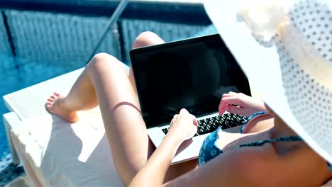 Mujer-en-traje-de-baño-y-sol-sombrero-escribir-texto-usando-laptop-tumbado-en-la-tumbona-junto-a-la-piscina