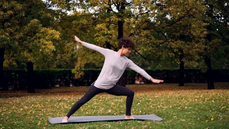 Hermosa-chica-con-pelo-rizado-corto-es-haciendo-yoga-al-aire-libre-permanente-en-pose-de-Guerrero-y-estirar-el-cuerpo-y-las-piernas-durante-la-práctica-en-el-parque.-Árboles-y-la-hierba-son-visibles.