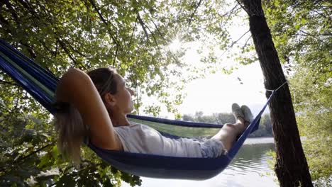 Mujer-joven-relajante-en-hamaca-por-el-río,-las-manos-detrás-de-la-cabeza-disfrutando-de-ambiente-verde-sereno.-Gente-viaje-relax-concepto-en-vacaciones