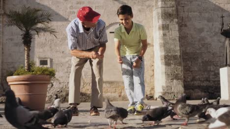 Grandparent-And-Grandson-Feeding-Pigeons-With-Bread-On-Vacations