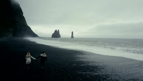 Vista-aérea-de-la-joven-pareja-caminando-en-la-playa-volcánica-negra-cerca-el-troll-del-pie-acantilados-en-Islandia