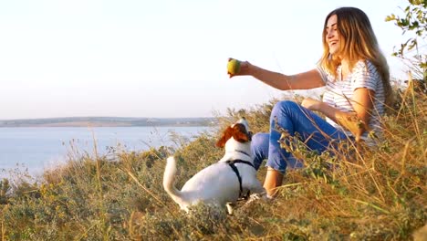 Mujer-atractiva-joven-jugando-con-un-perro-Jack-Russell-en-el-Prado-al-atardecer-con-el-fondo-del-mar.-cámara-lenta