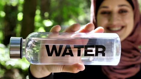 Portrait-of-a-cute-young-girl-in-a-hijab-with-a-bottle-of-water-in-her-hands,-smiling,-looking-at-the-camera,-park-in-the-background,-focus-pull-50-fps