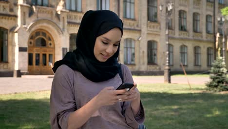 Junge-süße-muslimisches-Mädchen-Hijab-ist-Nachricht-auf-Smartphone-tagsüber-im-Sommer,-die-Eingabe-kichern,-aufbauend-auf-Hintergrund,-Religiuos-Konzept,-Kommunikations-Konzept