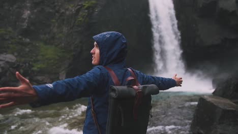 Back-view-shot-of-young-man-rising-hands-near-the-powerful-waterfall-in-Iceland,-cinematic-shot