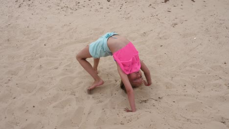 Young-acrobatic-girl-doing-gymnastic-bridge-and-somersaults-on-sand