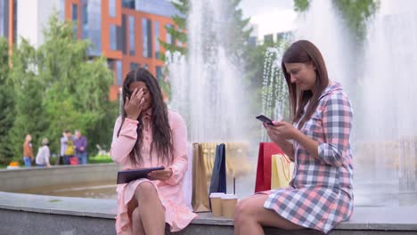 Dos-chicas-después-de-compras-están-sentados-en-el-parque-cerca-de-la-fuente-usando-un-teléfono-y-un-tablet.-4K