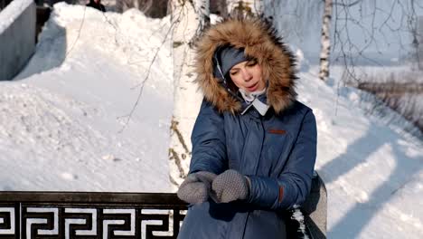 Woman-in-blue-down-jacket-with-fur-hood-calling-her-cellphone-in-winter-Park-and-put-on-her-mitten.