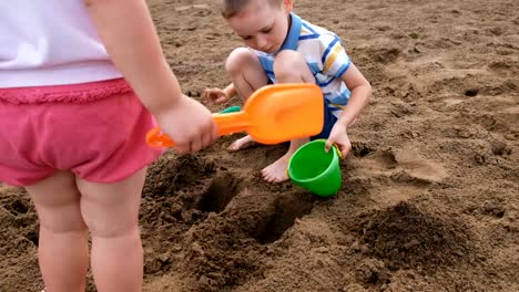 Kleiner-Junge-gießt-Wasser-aus-einem-Eimer-in-ein-Loch-in-den-Sand.-Kinder-spielen-am-Strand
