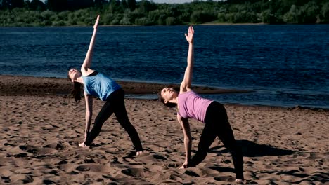 Zwei-Frau-Yoga-am-Sandstrand-am-Fluss-in-der-Stadt-zu-tun.-Blick-auf-die-schöne-Stadt-im-Sonnenaufgang.-Trikonasans-Utiha-darstellen.