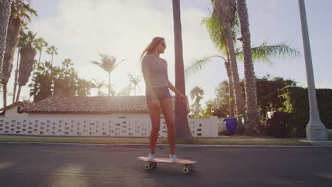 Attractive-adventurous-girl-skateboarding-down-palm-tree-lined-street-at-sunset