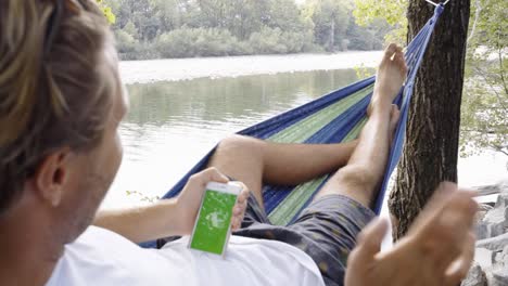 Hombre-joven-con-teléfono-móvil-en-hamaca-relajante-por-el-río-en-verano