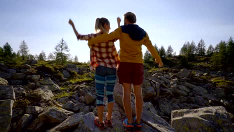 Young-couple-on-a-hike-surrounded-by-mountain-peaks-standing-arms-outstretched-in-front-of-the-sun,-Couple-hiking-arms-wide-open-freedom-and-achievement-concept