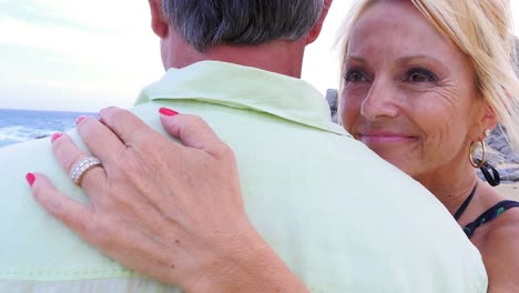 Close-up-of-an-older-woman-smiling-and-whispering-"I-love-you"-into-her-husband's-ear-at-the-beach