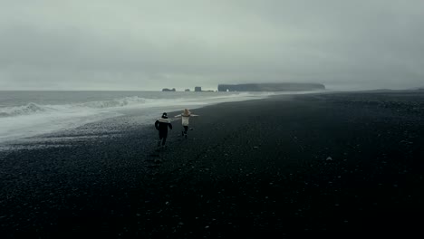 Copter-flies-after-young-couple-running-on-the-shore-of-the-sea-in-icelandic-sweater.-Black-volcanic-beach-in-Iceland