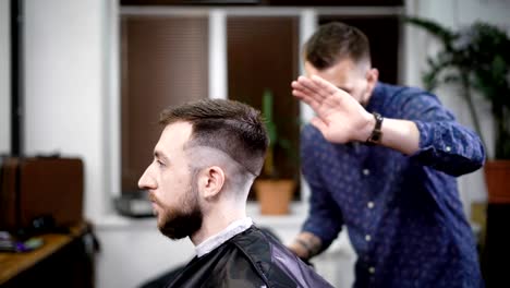 Young-barber-standing-and-making-stylish-haircut-of-attractive-man-with-clipper-in-barbershop.-Bearded-man-is-sitting-on-the-chair-with-black-protective-cape-against-the-mirror-and-looking-at-himself