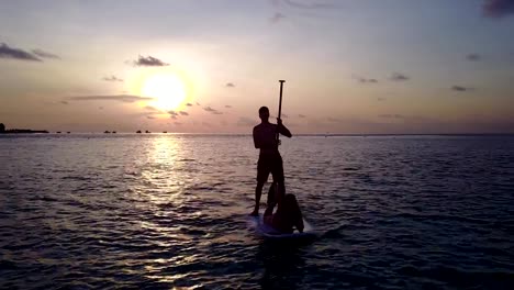 v04105-Aerial-flying-drone-view-of-Maldives-white-sandy-beach-2-people-young-couple-man-woman-paddleboard-rowing-sunset-sunrise-on-sunny-tropical-paradise-island-with-aqua-blue-sky-sea-water-ocean-4k