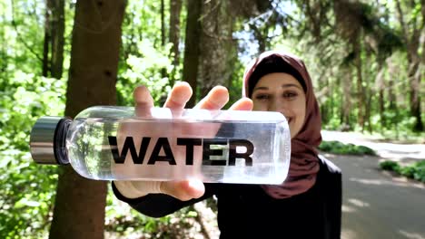 Portrait-of-a-cute-young-girl-in-a-hijab-with-a-bottle-of-water-in-her-hands,-smiling,-looking-at-the-camera,-park-in-the-background,-focus-pull-50-fps