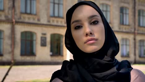 Young-self-confident-muslim-girl-is-watching-at-camera-in-daytime-in-summer-,-building-on-background,-religious-concept