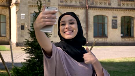 Joven-hermosa-chica-musulmana-en-hijab-es-hacer-selfie-en-su-smartphone,-mostrando-como-señal-durante-el-día-en-verano,-basándose-en-antecedentes,-concepto-de-epoca,-concepto-de-comunicación