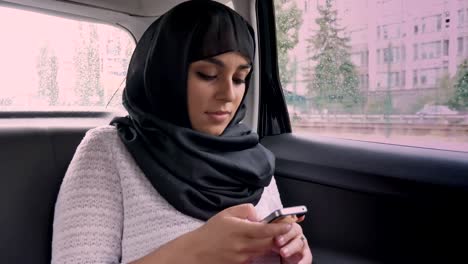 Young-muslim-woman-in-hijab-is-riding-in-car,-surfing-internrt-on-smartphone,-transport-concept,-communication-concept