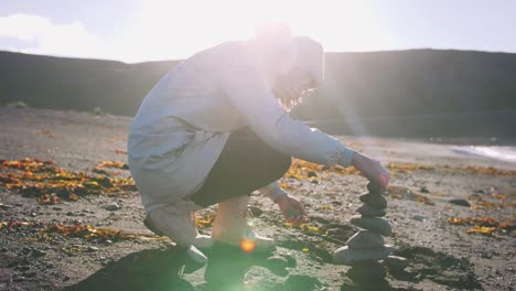 Beautiful-woman-stacking-stones-on-rock-at-sunny-black-beach-of-Iceland,-dolly,-slow-motion