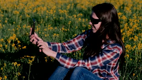 Woman-makes-a-selfie-on-the-phone-sitting-on-the-grass-among-the-yellow-flowers.