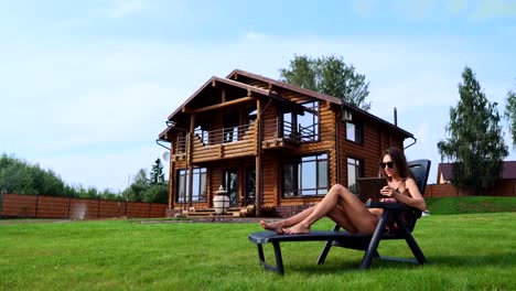 Hermosa-mujer-esbelta-acostado-en-una-hamaca-en-bañador-con-una-tableta-en-el-fondo-de-una-casa-rural-grande-en-gafas-de-sol