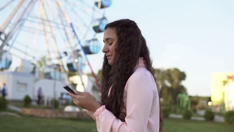 A-young-girl-walks-by-the-ferris-wheel-and-uses-a-smartphone.-4K