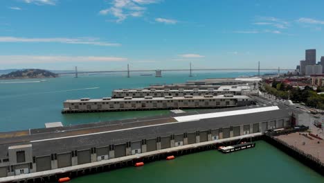 Drohne-geschossen-aufsteigend-über-eine-Gruppe-von-kommerziellen-Docks-in-San-Francisco-mit-Boote,-blauer-Himmel,-Wasser-und-der-San-Francisco-Oakland-Bay-Bridge-im-Blick