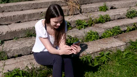 Woman-types-a-message-on-phone-sitting-in-park-and-smiling.