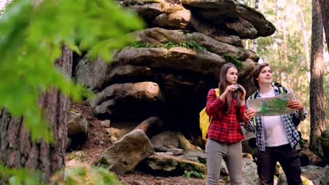 Lost-tourists-are-standing-in-forest-near-huge-rocks,-looking-at-map-then-through-binoculars-and-talking-discussing-way-to-their-destination-place.-People-and-nature-concept.