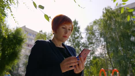 Serious-woman-with-red-hair-using-smartphone-outdoors-with-backlit,-low-angle