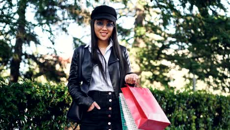 Portrait-of-smiling-Asian-girl-shopaholic-standing-outdoors-in-the-street-with-shopping-bags-and-looking-at-camera.-Green-trees-and-bushes-are-in-background.