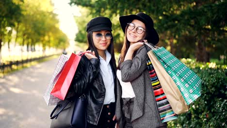 Portrait-of-beautiful-young-women-shoppers-standing-in-the-street-holding-paper-bags-and-smiling-looking-at-camera.-Youth-lifestyle-and-consumerism-concept.