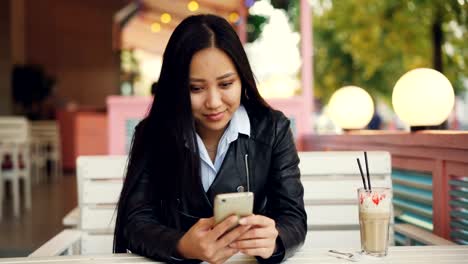 Mujer-asiática-moderna-es-utilizando-a-smartphone-mensajes-amigos-sentado-en-café-de-la-calle-en-día-ventoso-de-otoño-y-sonriendo.-Concepto-de-tecnología,-la-comunicación-y-la-gente.