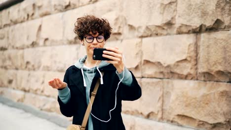 Stylish-young-lady-is-making-video-call-online,-talking-and-gesturing-using-smartphone-and-looking-at-screen-wearing-earphones.-Conversation,-technology-and-people-concept.