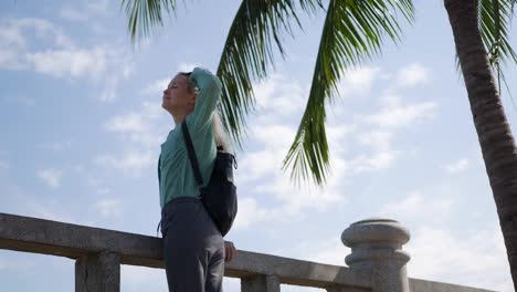 Joven-caucásica-feliz-con-cabello-largo-Rubio-en-gafas-de-sol-y-camiseta-verde-de-pie-y-sonriendo-junto-a-la-palmera-sobre-un-fondo-de-cielo-azul.-Concepto-de-viaje