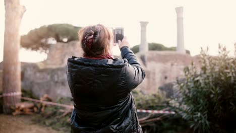 Back-view-of-happy-senior-smiling-Caucasian-woman-taking-a-smartphone-photo-of-ancient-ruins-in-Ostia,-Italy-on-vacation