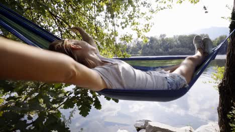 Mujer-joven-relajante-en-hamaca-por-el-río,-las-manos-detrás-de-la-cabeza-disfrutando-de-ambiente-verde-sereno.-Gente-viaje-relax-concepto-en-vacaciones