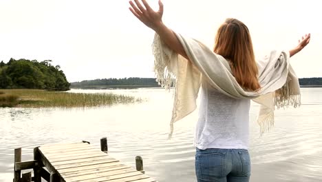 Young-woman-relaxes-on-lake-pier,-stands-arms-outstretched