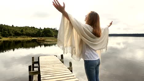 Young-woman-relaxes-on-lake-pier,-stands-arms-outstretched