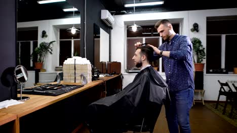 Joven-peluquero-en-ropa-informal-permanente-y-peinado-a-cliente-antes-de-cortar-su-pelo.-Hombre-sonriente-sentado-en-una-silla-cubierta-con-bata-negra-y-mirando-en-el-espejo-frente-a-la-peluquería