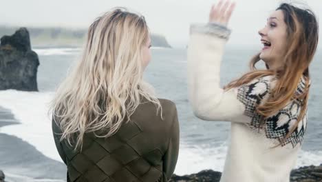 Dos-mujeres-viajan-de-pie-en-la-playa-negra,-llevar-los-pies-del-troll-rocas-en-Islandia-y-en-agua,-sonreír,-divertirse