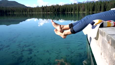 Woman's-feet-dangle-from-wooden-pier,-above-lake