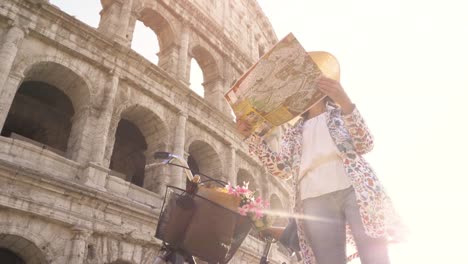 Beautiful-young-woman-in-colorful-fashion-dress-with-bike-reading-map-in-front-of-colosseum-in-Rome-at-sunset-with-happy-attractive-tourist-girl-with-straw-hat-looking-for-directions-ground-shot-dolly-moving-camera