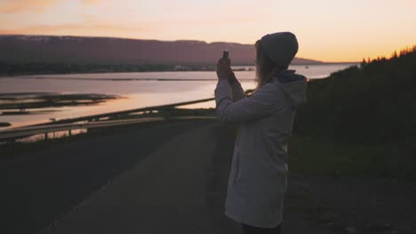 Silueta-de-una-mujer-sosteniendo-un-smartphone-tomando-fotos-fuera-en-la-carretera-de-montaña-durante-la-puesta-de-sol