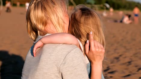 Beautiful-blonde-mom-and-daughter-cuddling-on-the-beach-at-sunset.