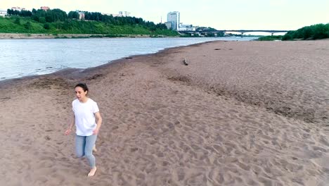 Mujer-de-correr-a-lo-largo-de-la-playa-de-arena-de-río-al-atardecer.-Vista-hermosa-ciudad.