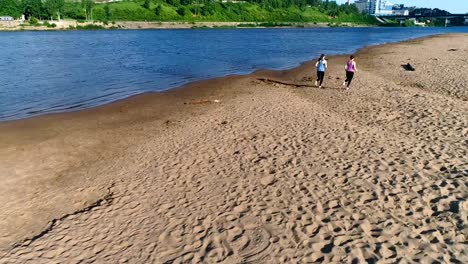 Dos-mujeres-corriendo-a-lo-largo-de-la-playa-de-arena-de-río-al-atardecer.-Vista-hermosa-ciudad.